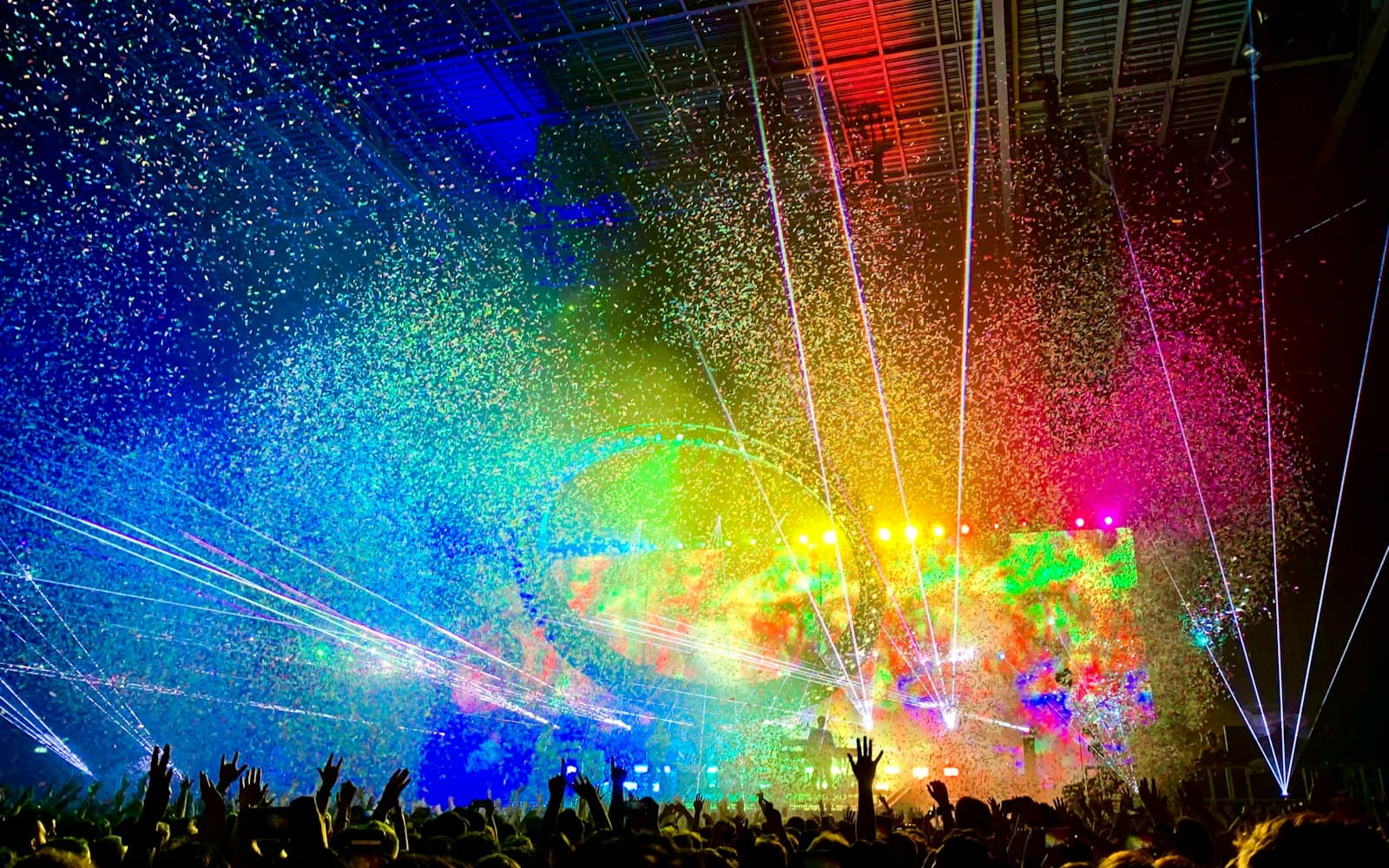 Confetti and laser beams filling an arena above a crowd with raised hands during a concert finale