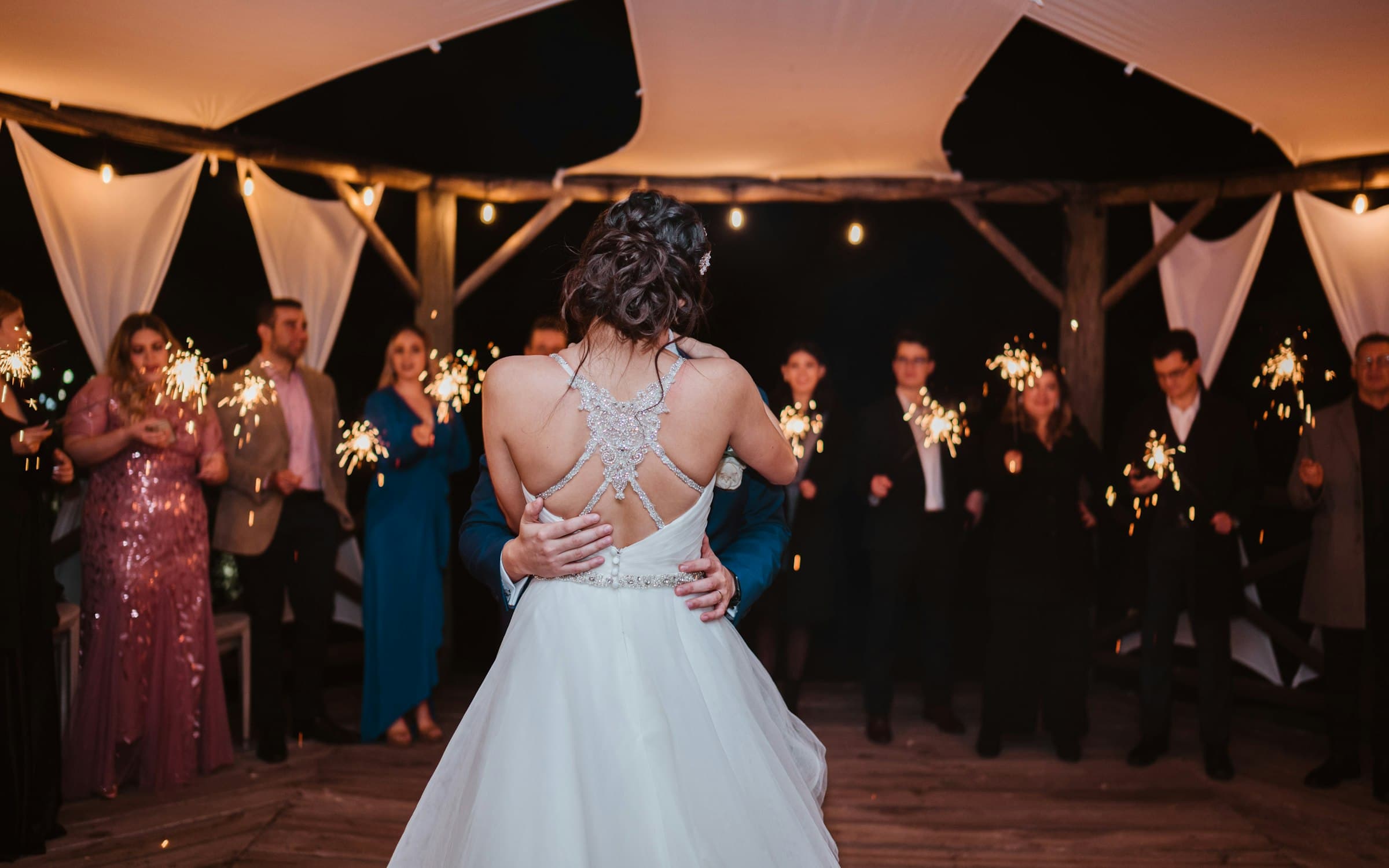 Couple sharing a first dance on a covered wooden deck at night while guests hold lit sparklers, the kind of moment couples now plan cold sparks around