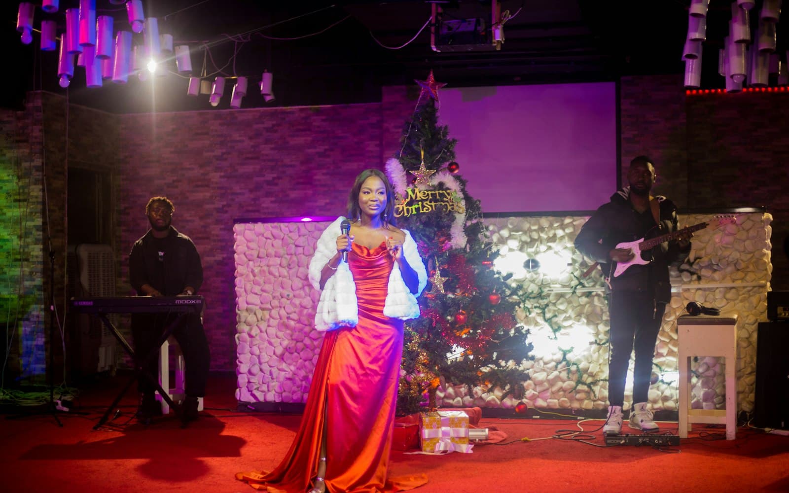 Vocalist in a red satin gown singing with a keyboardist and electric guitarist on a stage beside a decorated Christmas tree, a live band format for a company holiday party