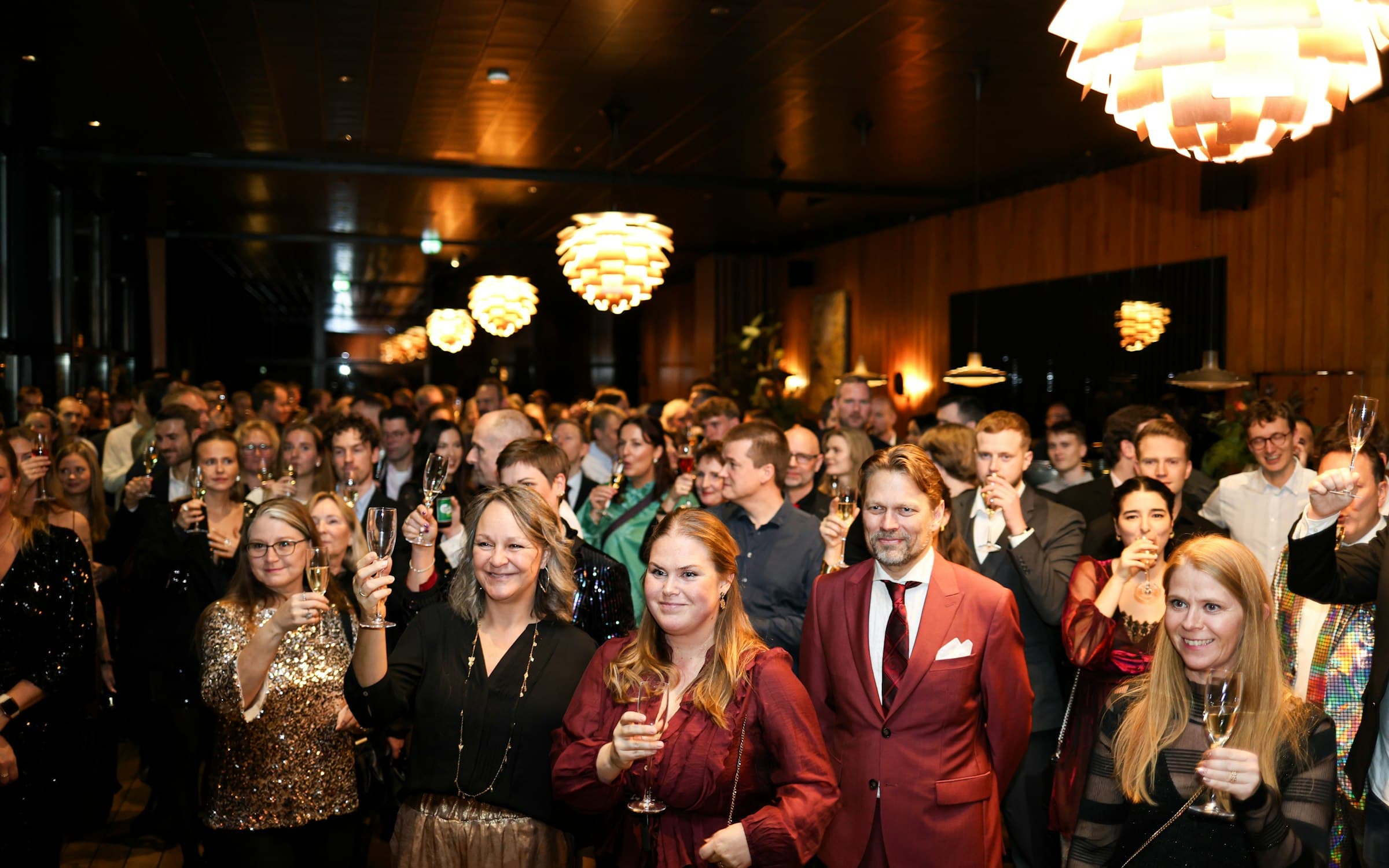 Guests in sequins and festive red attire raise champagne glasses during a toast at a crowded indoor celebration, the moment corporate holiday party entertainment builds toward
