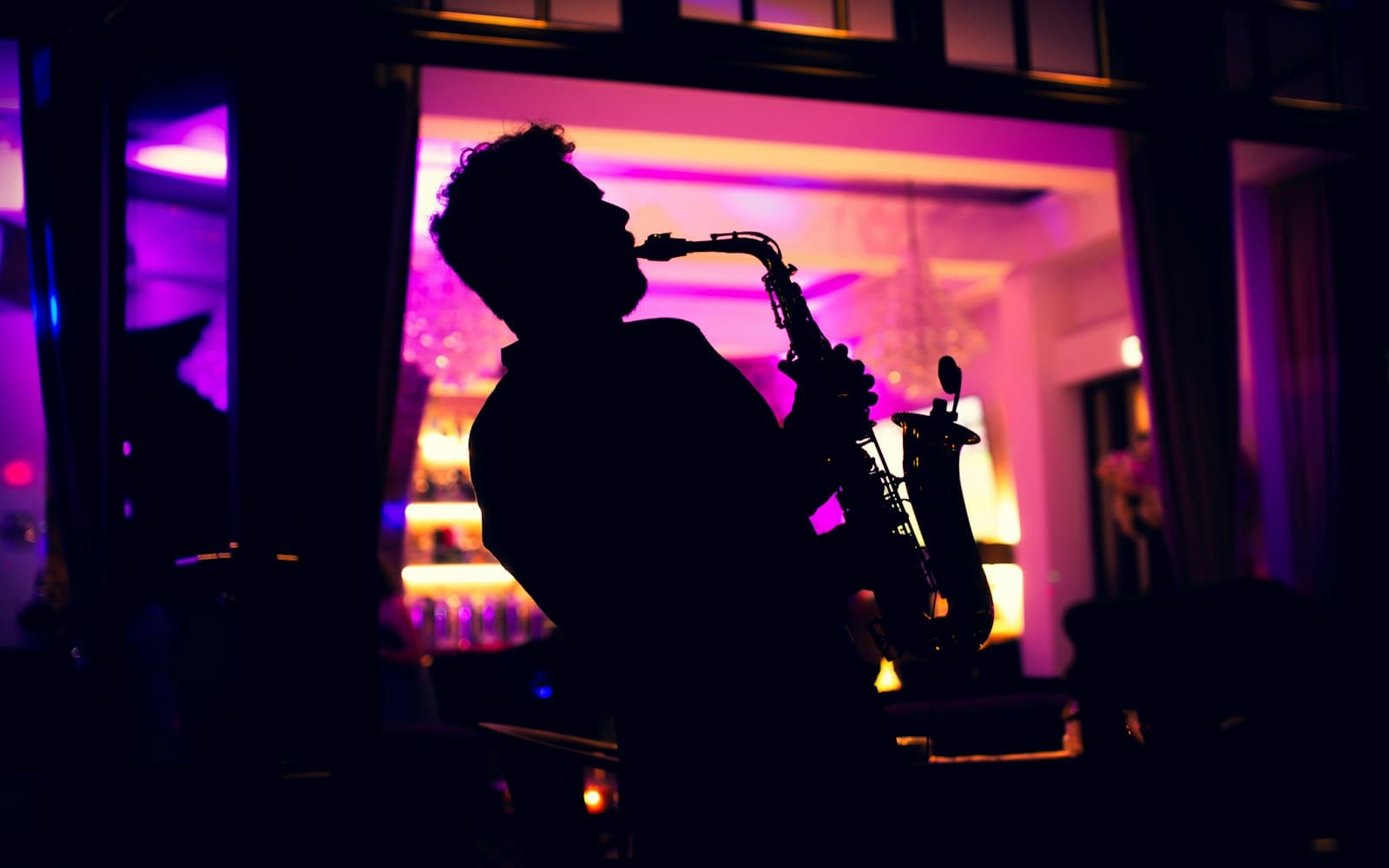 Silhouette of a saxophonist playing in front of a magenta-lit bar, a live sax feature that pairs well with a DJ at a holiday party