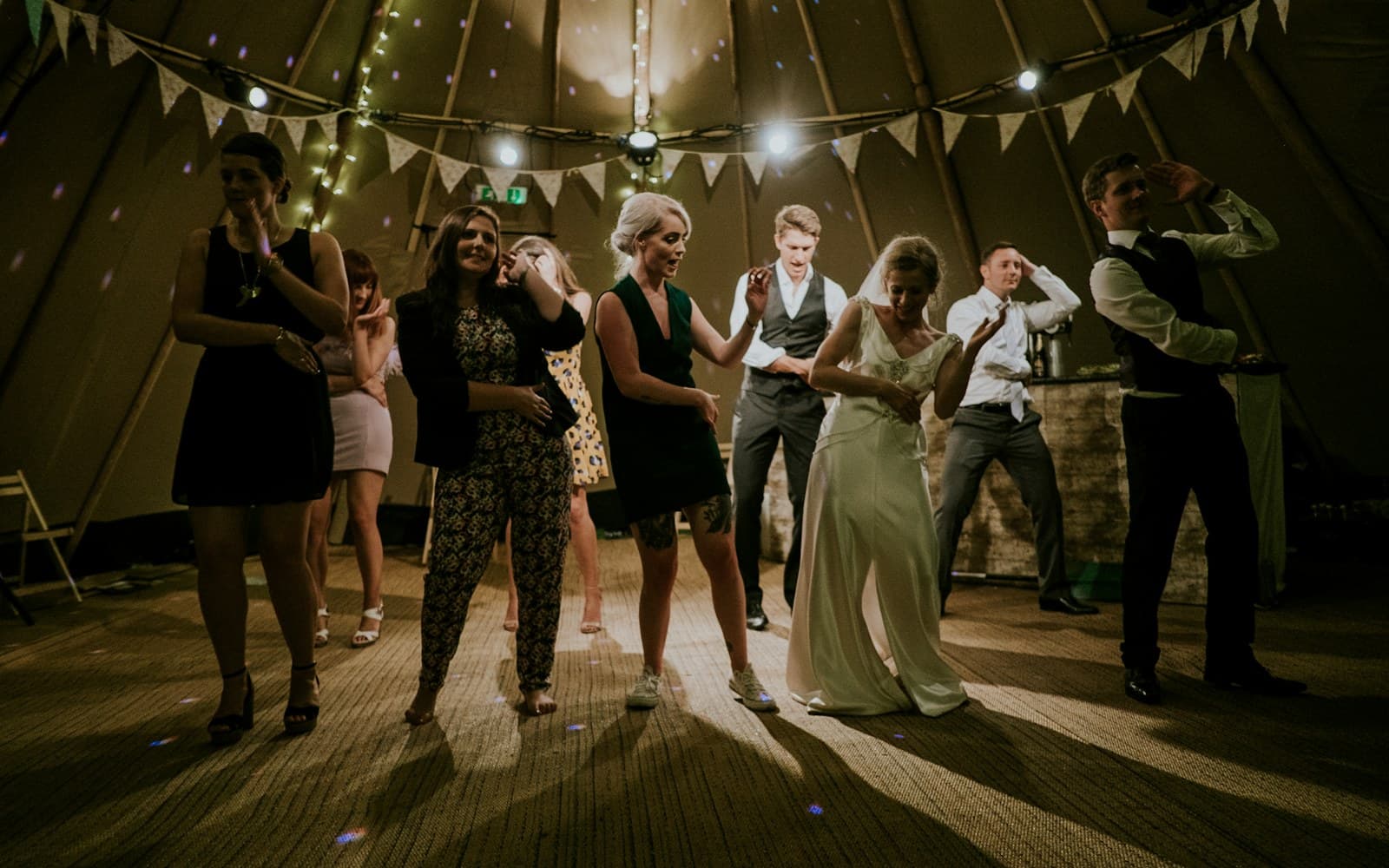 Wedding guests filling the dance floor under string lights and bunting — the full floor a live band or DJ is ultimately hired to deliver