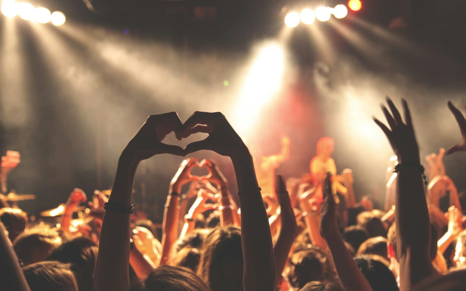 Crowd silhouetted under warm stage lights and atmospheric haze at an outdoor live event, with hands raised