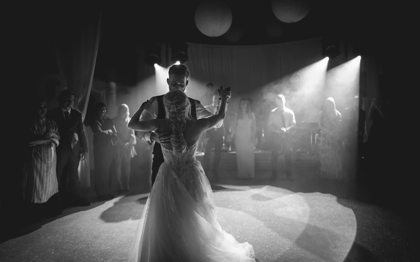 Black-and-white photo of a couple's first dance under stage spotlights and light haze, with guests watching from the edge of the dance floor