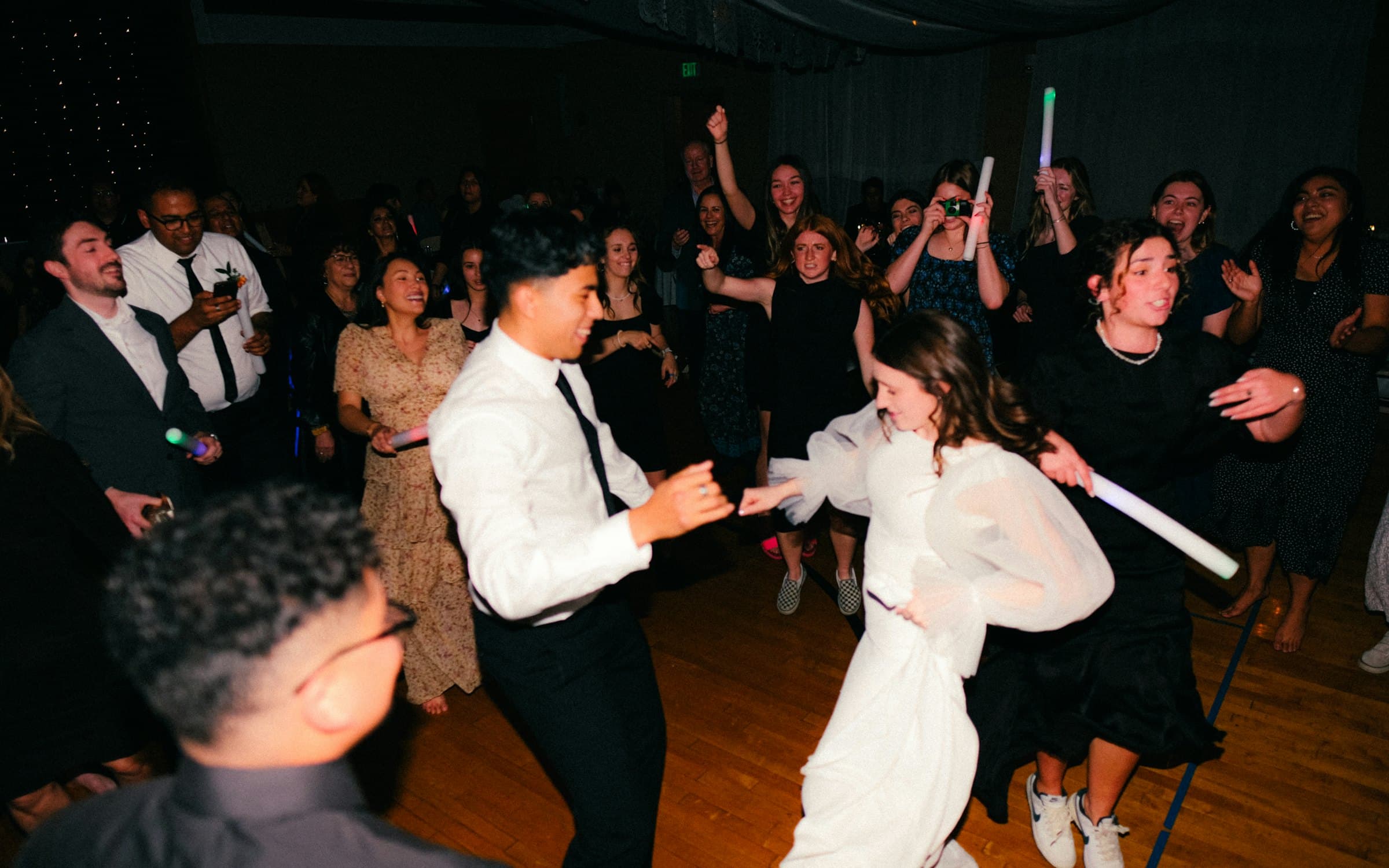 Bride and groom dancing in the middle of a packed wedding reception dance floor as guests cheer and wave glow sticks, the moment a good wedding DJ builds toward