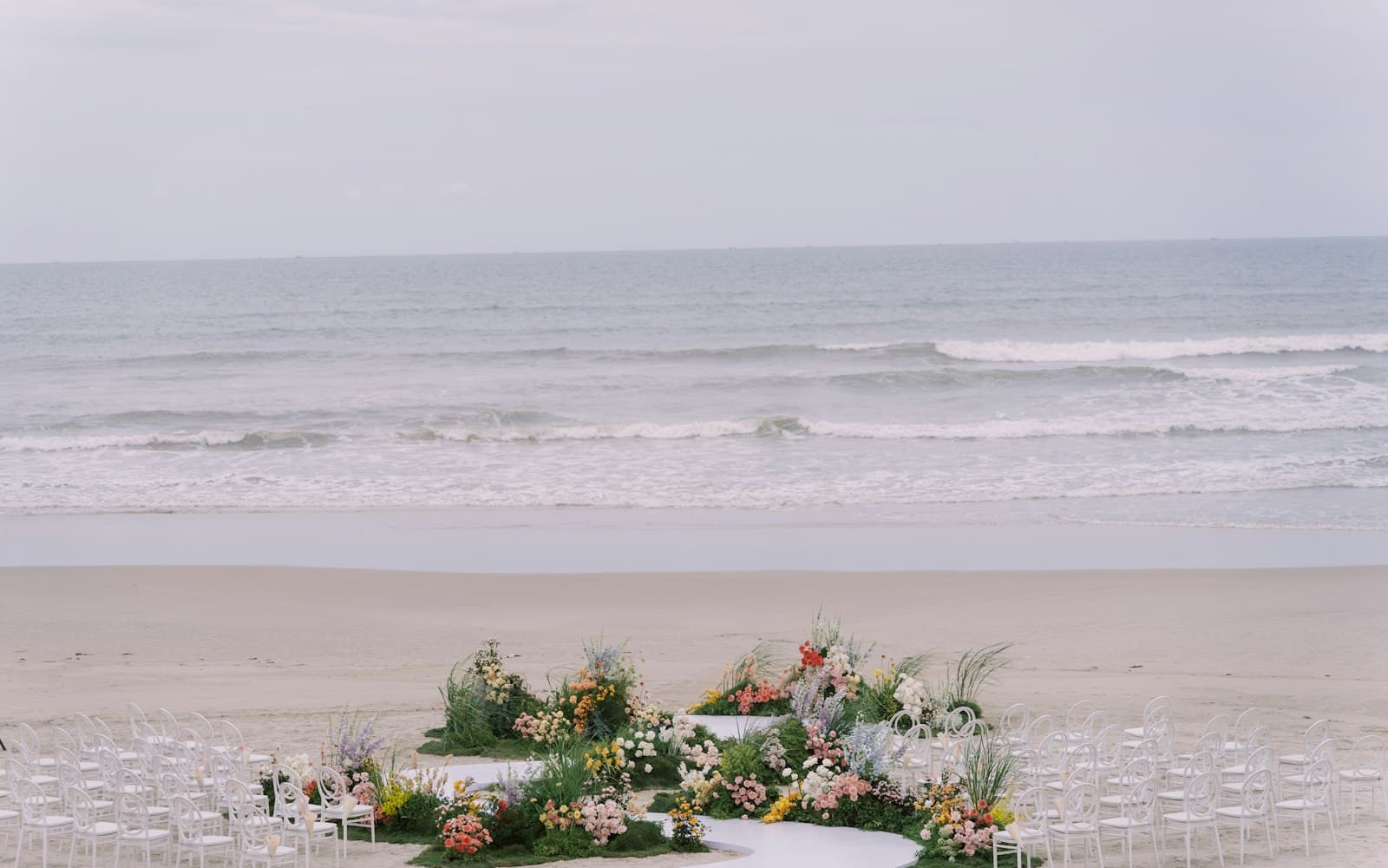 Beach wedding ceremony set up on the sand, with white chairs curved around a winding aisle of colorful flowers and waves breaking behind