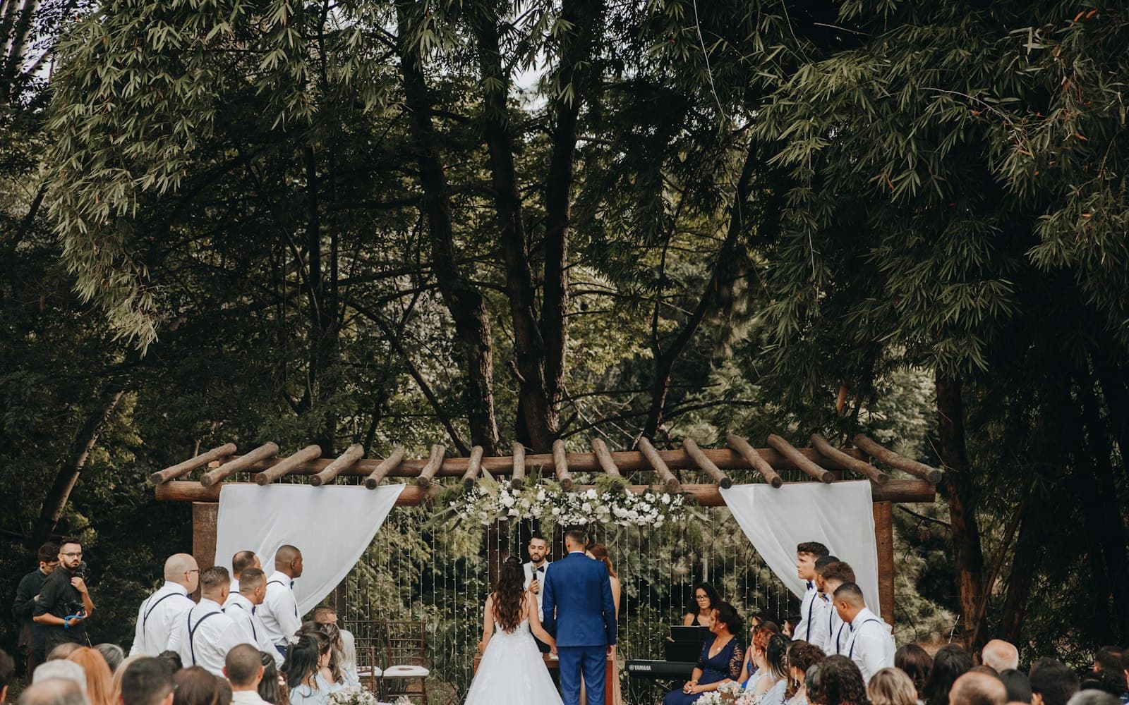 Outdoor wedding ceremony under a wooden pergola surrounded by tall trees, with the officiant speaking into a handheld microphone and a keyboardist seated beside the altar