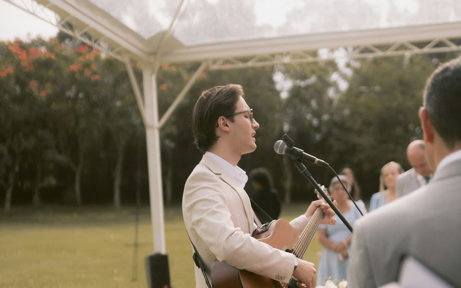 Vocalist-guitarist in a cream suit singing into a microphone during an outdoor wedding ceremony under a clear tent, with guests looking on