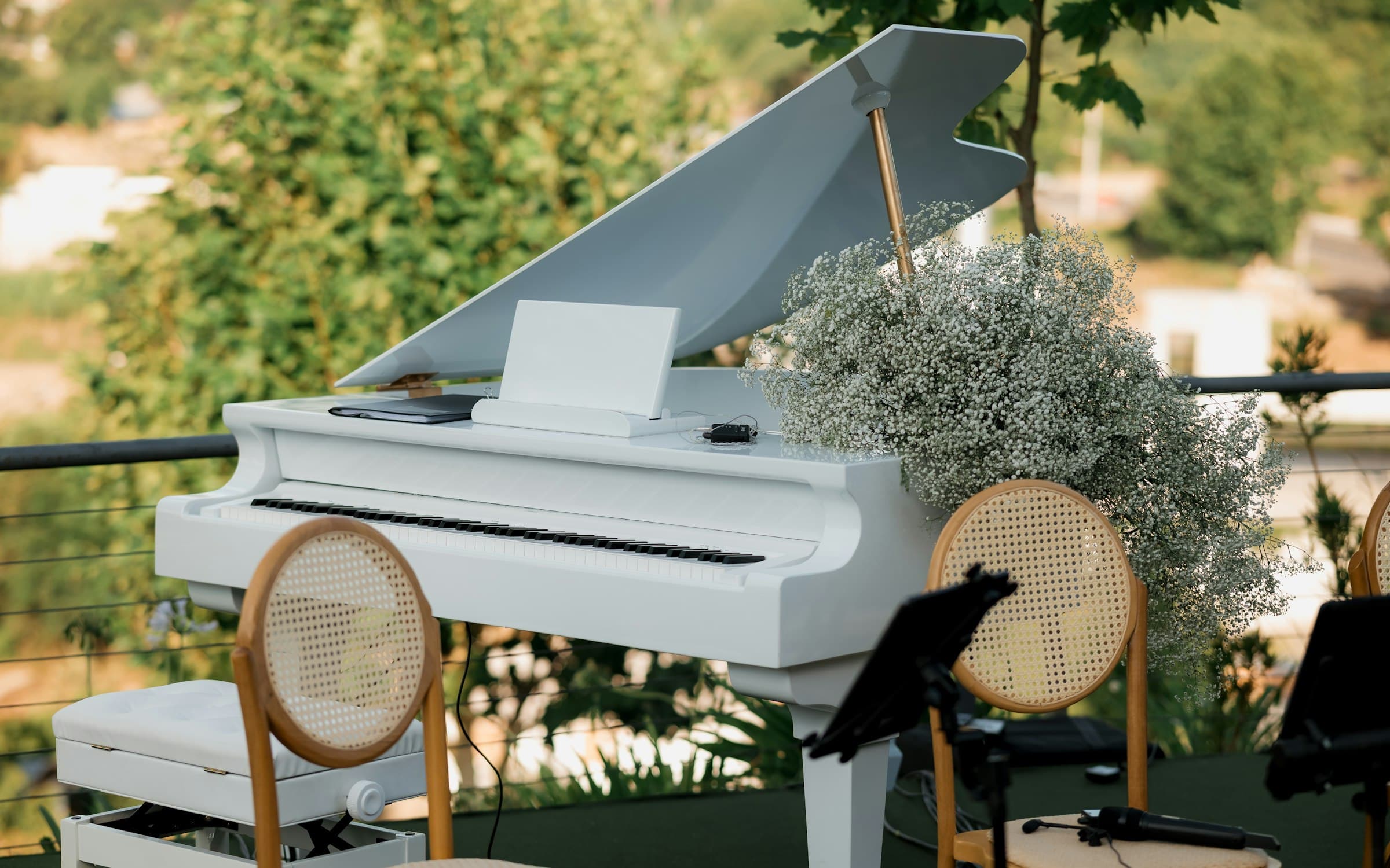 White grand piano dressed with baby's breath beside cane-back chairs and music stands, set up on an outdoor terrace for live wedding ceremony musicians