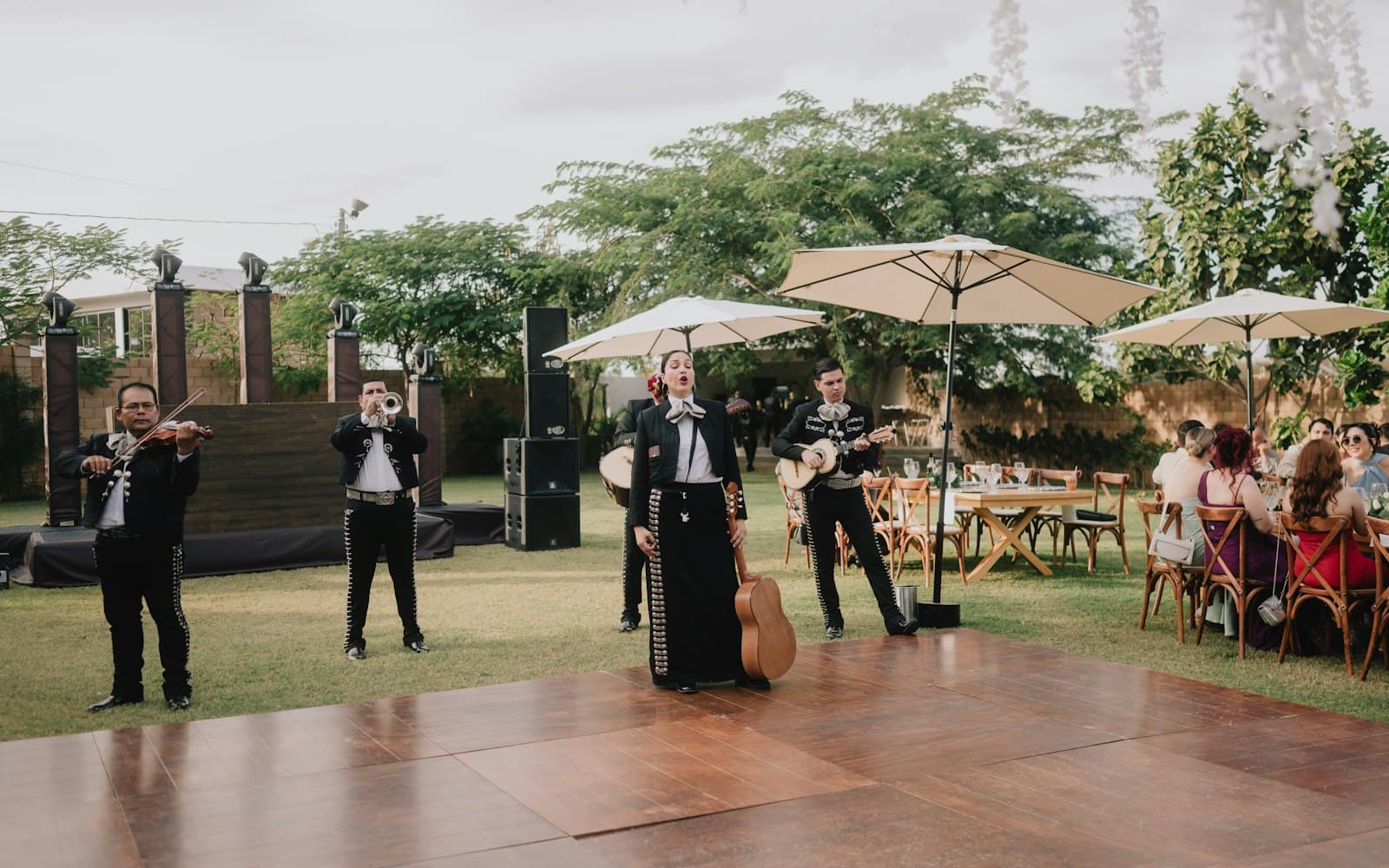 Mariachi ensemble with violin, trumpet, and guitars performing on a wooden dance floor at an outdoor wedding celebration as guests sit at tables under patio umbrellas